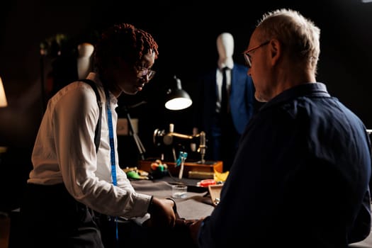 Attentive african american apprentice carefully listening to master dressmaker guiding her with cutting fabric. Couturiers manufacturing upcoming bespoken fashion collection