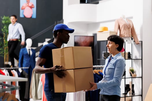 African american deliveryman holding carton packages, discussing shipping detalis with employee in shopping centre. Courier with blue uniform working with fashion store. Stylish boutique concept