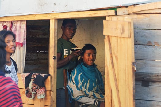 Ranohira, Madagascar - April 29, 2019: Unknown local boy getting his haircut from a friend at shop back room made from wooden boards. People in this part of Africa are poor, but cheerful.