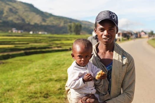 Manandoana, Madagascar - April 26, 2019: Unknown Malagasy man holding child with piece of food on his hand, rice fields in background on sunny day. People in this part of Africa are poor, but cheerful