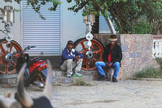 Ranohira, Madagascar - April 29, 2019: Two unknown local men resting on stone house porch in the evening, orange wheel pump next to them. People in this part of Africa are poor, but cheerful