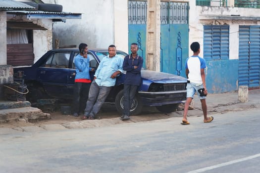 Ranohira, Madagascar - April 29, 2019: Three unknown local men leaning on old broken car in evening, resting and chatting. Another man walking by. people are poor here, but mobile phones are common.