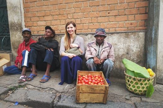 Antananarivo, Madagascar - April 24, 2019: Young caucasian woman sitting with local fruit sellers at Antananarivo high street. Despite poor living conditions people here are friendly and cheerful