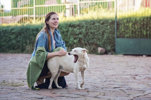 Candid portrait of young woman kneeling outside on stone ground, covered with blanket, holding stray dog yawning next to her