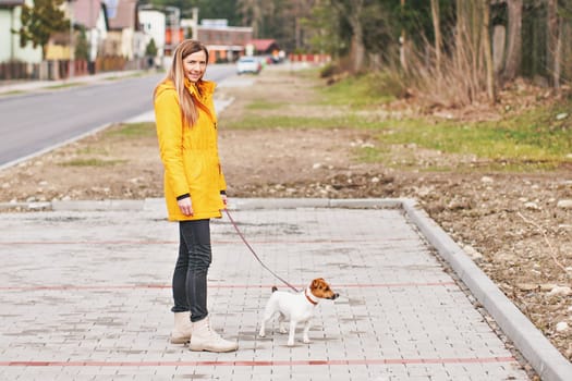 Young woman in yellow jacket, smiling and looking into camera, with Jack Russell terrier dog on a pink leash, blurred houses in background