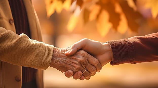 tender moment where a young person's hand is clasped in a warm handshake with an elderly individual, The backdrop of golden autumn leaves adds a sense of warmth and seasonal beauty to this touching scene of connection across generations