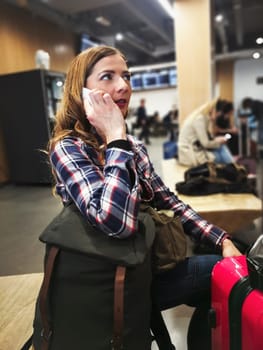 Young woman in shirt waiting for her transfer, with her backpack and luggage near her, talking on the phone, blurred station in background