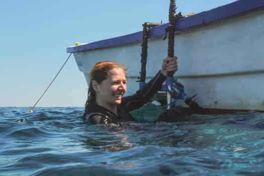 Young woman in neoprene, smiling, mask already removed, getting to boat from sea after her dive.