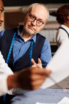 Close up of master tailor checking aspiring fashion design apprentice sartorial garment sketches in atelier workspace. Couturiers working on bespoken client customization comission