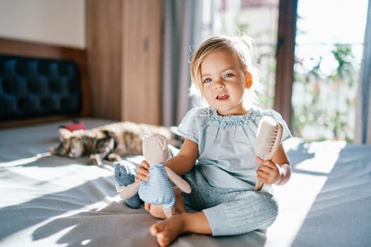 Little smiling girl sitting on the bed with a teddy bear and a comb in her hands. High quality photo