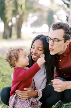 Dad blows on a dandelion in the hands of a little girl standing next to squatting mom. High quality photo