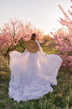 Woman blooming peach orchard. Against the backdrop of a picturesque peach orchard, a woman in a long white dress and hat enjoys a peaceful walk in the park, surrounded by the beauty of nature