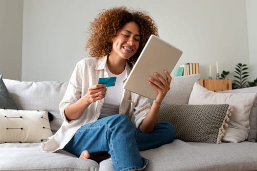 Happy and smiling young Multiracial hispanic woman online shopping using digital tablet and credit card sitting on the couch at home. E-commerce concept.