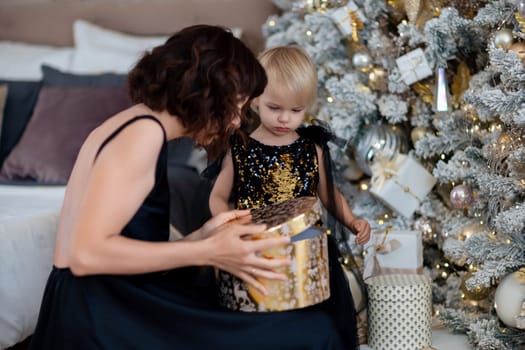 Mother daughter 2 years old Christmas tree. Both are dressed in black dresses, the mother holds the girl in her arms and both look at each other. The family celebrates Christmas