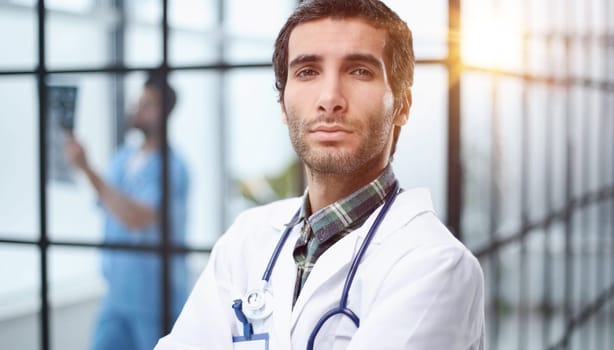 male doctor wearing white coat, stethoscope looking at camera standing with arms crossed in medical office.