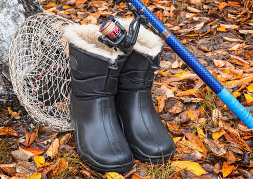 On earth on a background of autumn leaves are the items for fishing: landing net, spinning and warm waterproof boots.