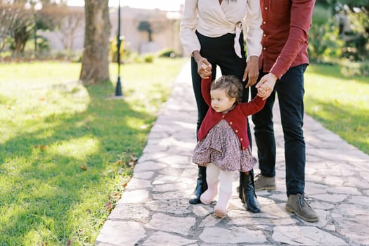 Little girl walks along a paved path in the garden holding hands of mom and dad. Cropped. High quality photo