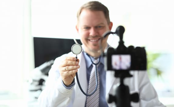 Focus on male hand holding and showing stethoscope to camera. Smiling doctor wearing white medical gown. Content maker smiling to camcorder. Blogging concept