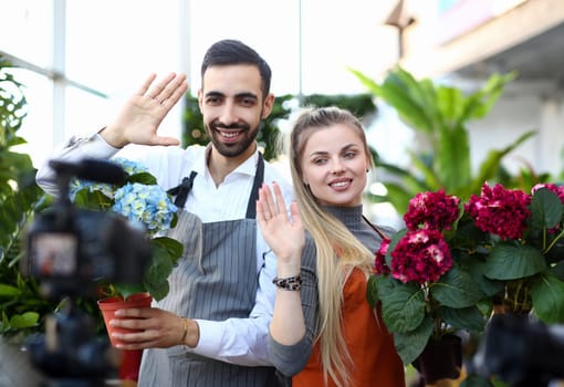 Smiling Vlogger Recording Hydrangea in Flowerpot. Woman and Man with Blooming Red and Blue Hortensia Waving to Camcorder. Domestic Plant Vlog for Gardeners. Cheerful Florist in Botanic Store