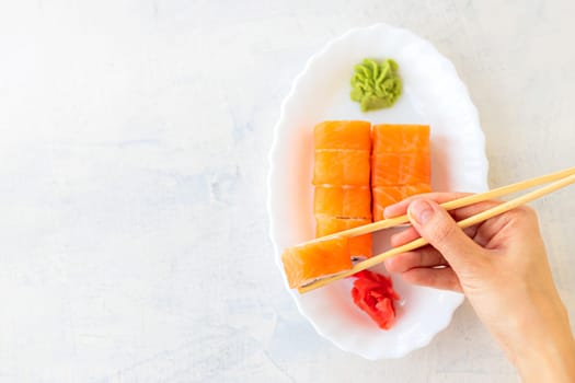 top view on female hands holding Japanese sushi or Philadelphia roll with wooden chopsticks on a light textured background, soft focus, copy space, flat lay,