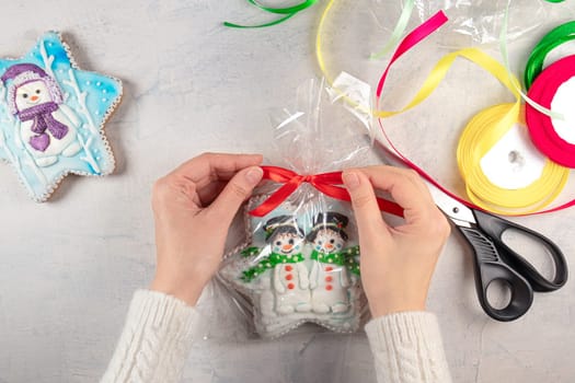 female hands in a white sweater tie a red bow on a package of gingerbread cookies with snowmen on the background of a light table, multi-colored ribbons and scissors. concept for self-made Christmas or New Year gifts.