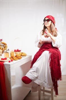 A fashionable modern girl in stylized folk clothes at table with a samovar, bagels and tea for the Orthodox holiday of Maslenitsa and Easter. Funny photo shoot for a young woman