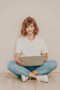 A brunette sits on the floor with a laptop on a beige wall background. She is wearing a white T-shirt, jeans and white sneakers