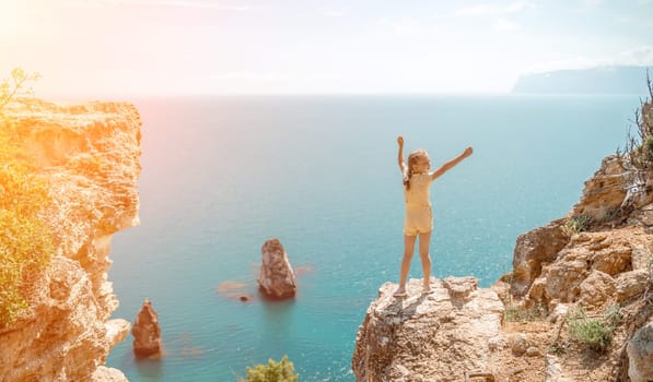 Happy girl stands on a rock high above the sea, wearing a yellow jumpsuit and sporting braided hair, depicting the idea of a summer vacation by the sea