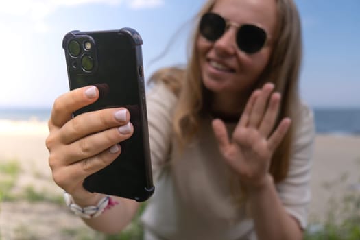 Young woman in sunglasses with shell bracelets holding smartphone. Influencer Using mobile phone for video call selfie waving on the beach seacoast. Holiday vacation advertisement concept. Social media