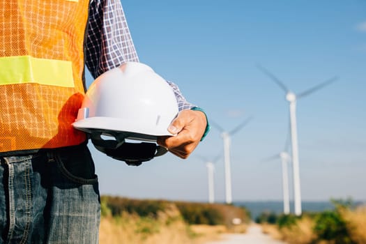 Engineer stands at wind turbine field holding safety helmet. A symbol of renewable success innovation in reducing global warming. Demonstrates leadership commitment to safety in industry.