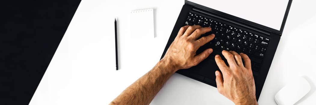 The man working at home on the remote. A black-and-white workplace with a laptop, alarm clock, notebook and men's hands.