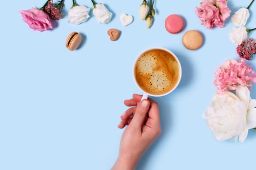 Blue background with macaroons and a Cup of coffee in a hand surrounded by peonies and roses. Top view with space for your text.