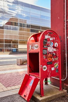 Vintage red telephone booth adorned with graffiti, standing against a modern building in Muncie, Indiana - a symbol of times changing in urban life.