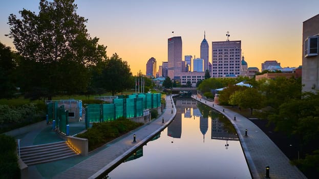 Sunrise over Indianapolis: Aerial view of urban skyline reflecting on canal waters, taken from a drone during golden hour, highlighting city's green park and modern architecture.