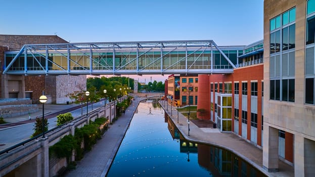 Twilight over Indianapolis: Aerial view of a glowing pedestrian bridge over a tranquil canal, captured by DJI Mavic 3 drone, exhibiting a harmony of modern architecture and waterway.