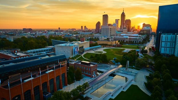 Golden hour cityscape of Indianapolis, displaying a vibrant mix of modern and traditional architecture, with a serene urban park in the foreground, captured by a drone.