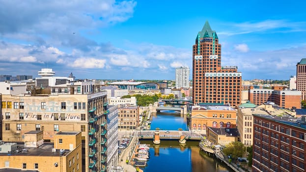 Aerial view of diverse architecture in Milwaukee, from historic to modern buildings, under a clear blue sky, with a prominent green-roofed structure overlooking the bustling cityscape and riverside leisure activities, captured by a DJI Mavic 3 drone.