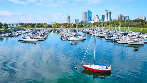 Sailboats and yachts docked in vibrant Milwaukee marina, with a standout red-hulled sailboat, under a clear blue sky - a blend of urban cityscape and peaceful lakeside leisure.