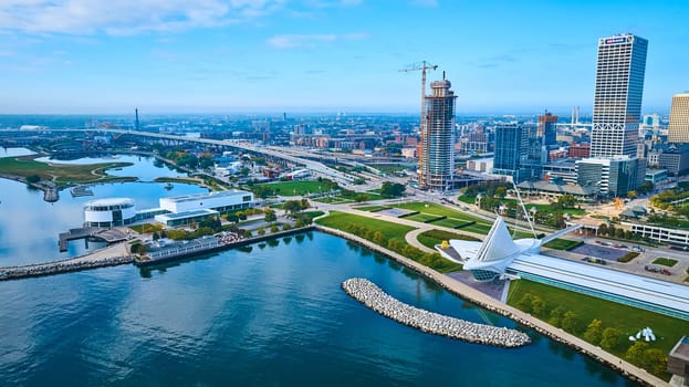 Aerial view of Milwaukee's vibrant waterfront cityscape featuring a modern suspension bridge, the glassy Quadracci Pavilion and a rising skyscraper under construction, depicting urban growth and harmony with nature.