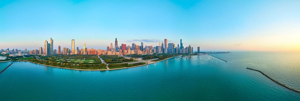 Sunrise over Chicago's skyline showcasing a mix of modern and historic architecture, with Lake Michigan's serene waters and lush green parks captured in a 2023 aerial panorama taken by a DJI Mavic 3 drone.