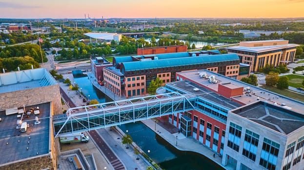 Golden hour aerial view of modern Indianapolis architecture, showcasing a serene water canal with pedestrian bridge and green urban spaces