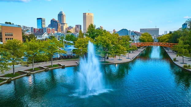 Sunlit water fountain in vibrant urban park with autumnal trees and modern city skyline, captured by drone in Indianapolis, 2023