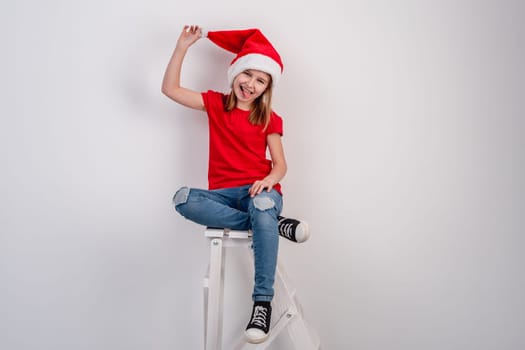 Little Girl In Red T-Shirt And Santa Hat Sits On White Stairs In Room For A Christmas Photo On A White Background