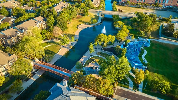 Aerial View of Bustling Suburban Park at Golden Hour in Indianapolis, Featuring River, Playground and Pedestrian Bridge
