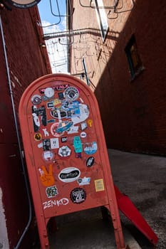 Sticker-adorned sandwich board in gritty alleyway, a striking depiction of urban art culture in Muncie, Indiana, 2023