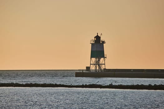 Image of Sunrise behind lighthouse at dawn with soft golden sky and Lake Michigan water in ocean endlessness