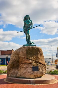 Bronze statue of a Native American warrior in traditional attire, standing on a boulder in downtown Muncie, Indiana, 2023, symbolizing strength and vigilance