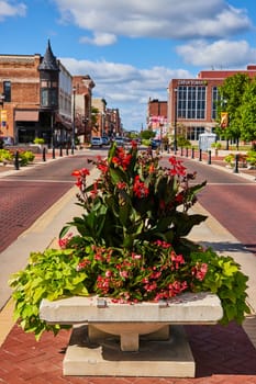 Vibrant urban street scene in Muncie, Indiana, showcasing a pedestrian-friendly sidewalk with a lush flower planter and diverse architecture under a sunny sky in 2023.