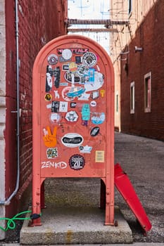 Vintage red mailbox, covered in eclectic stickers, standing in a narrow urban alleyway in downtown Muncie, Indiana, symbolizing community and artistic expression.
