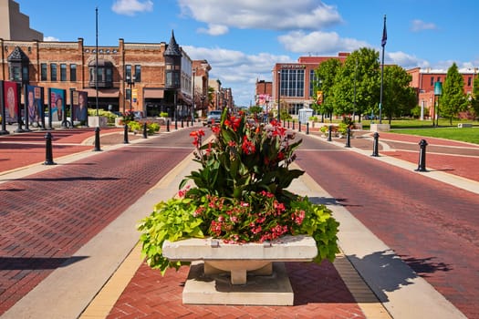 Vibrant downtown scene in Muncie, Indiana, 2023, showcasing a historic brick street lined with colorful banners and lush planters, under a tranquil blue sky.